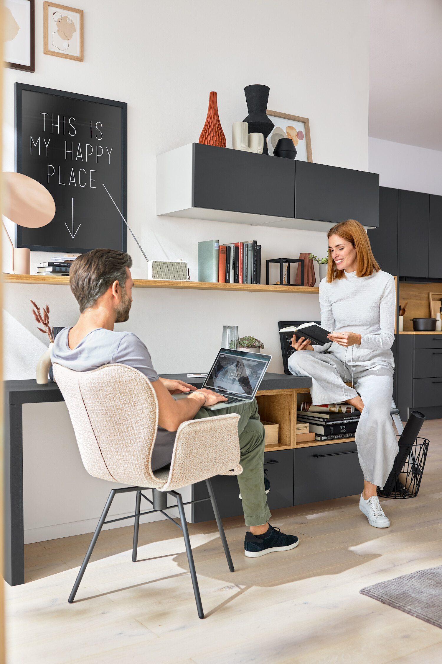 Ein Mann sitzt am Tisch mit seinem Laptop auf den Knien. Eine Frau sitzt ihm gegenüber auf dem Schrank und hat ein Buch aufgeschlagen.