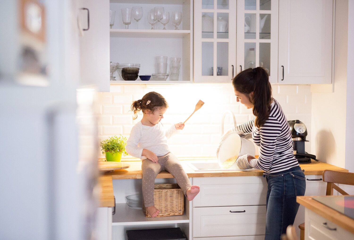 Blick in Landhausküche: ein Kind sitzt auf dem Schrank und hat einen Kochlöffel in der Hand, die Mutter gießt heißes Wasser in die Spühle.