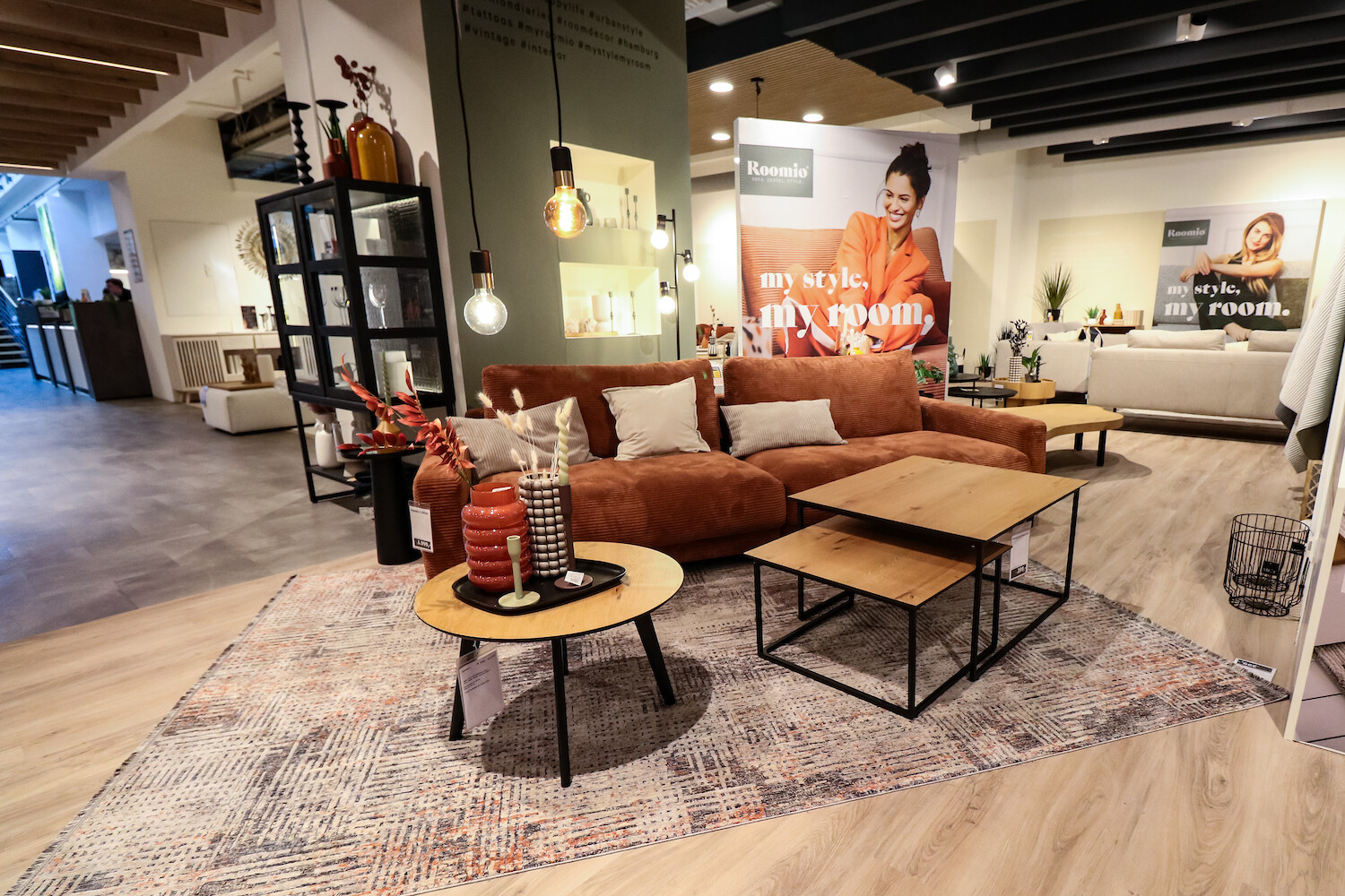 Blick in Ausstellungsraum auf Wohnzimmer mit Couch, rundem Beistelltisch und eckigem Couchtisch und hängenden Glühlampen.