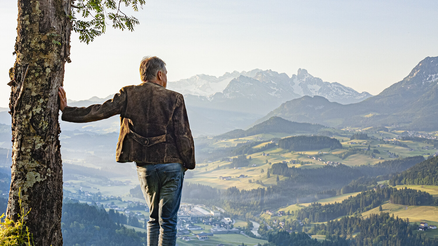 Ein Mann stützt sich an einem Baum ab und schaut auf ein Tal, im Hintergrund sind hohe Berge zu sehen.
