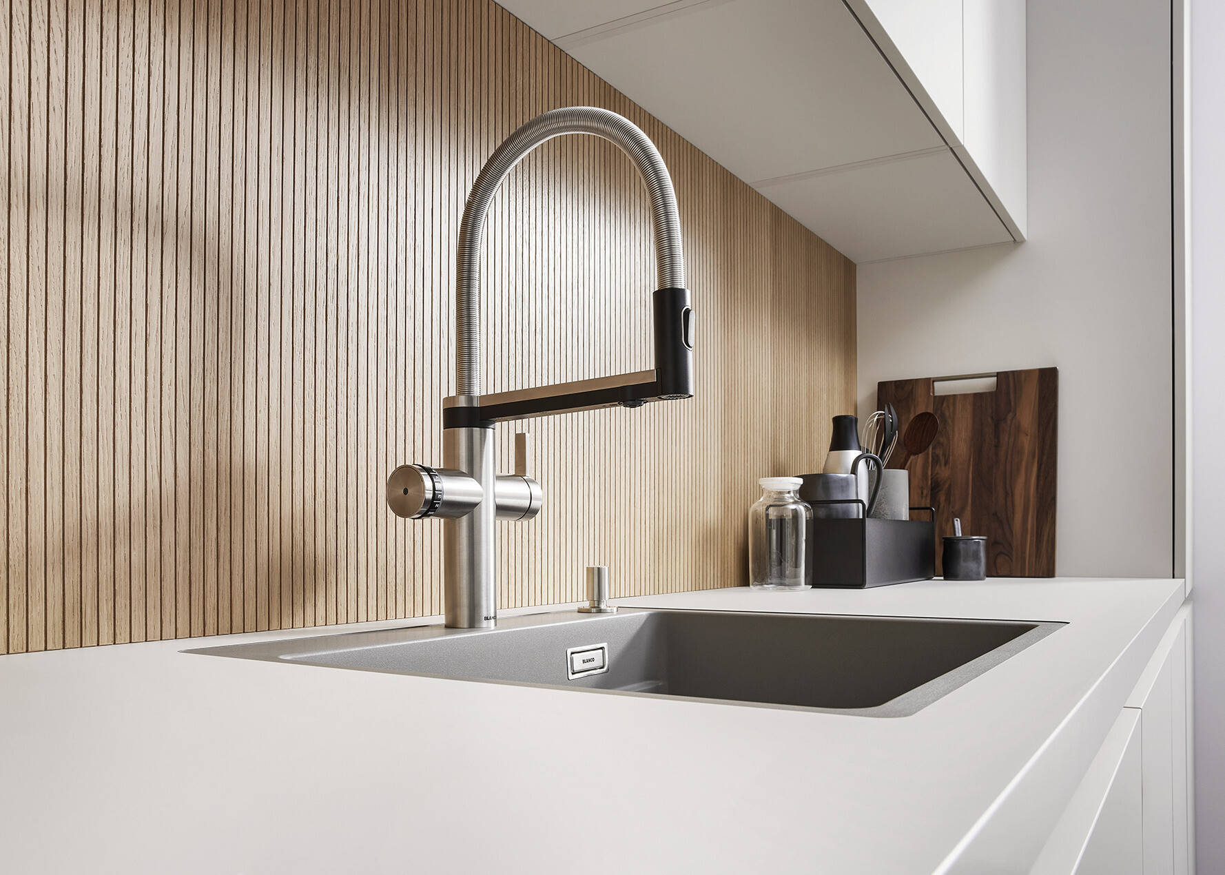 Zu sehen ist ein laufender Wasserhahn auf einer Küchenzeile mit einem Spülbecken. Zudem sieht man eine Kanne Tee, Zucker und Kuchen.